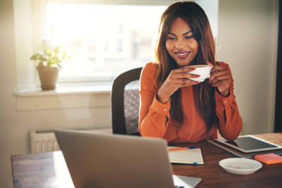 A woman enjoys a coffee at her desk as she watches a webinar on her laptop